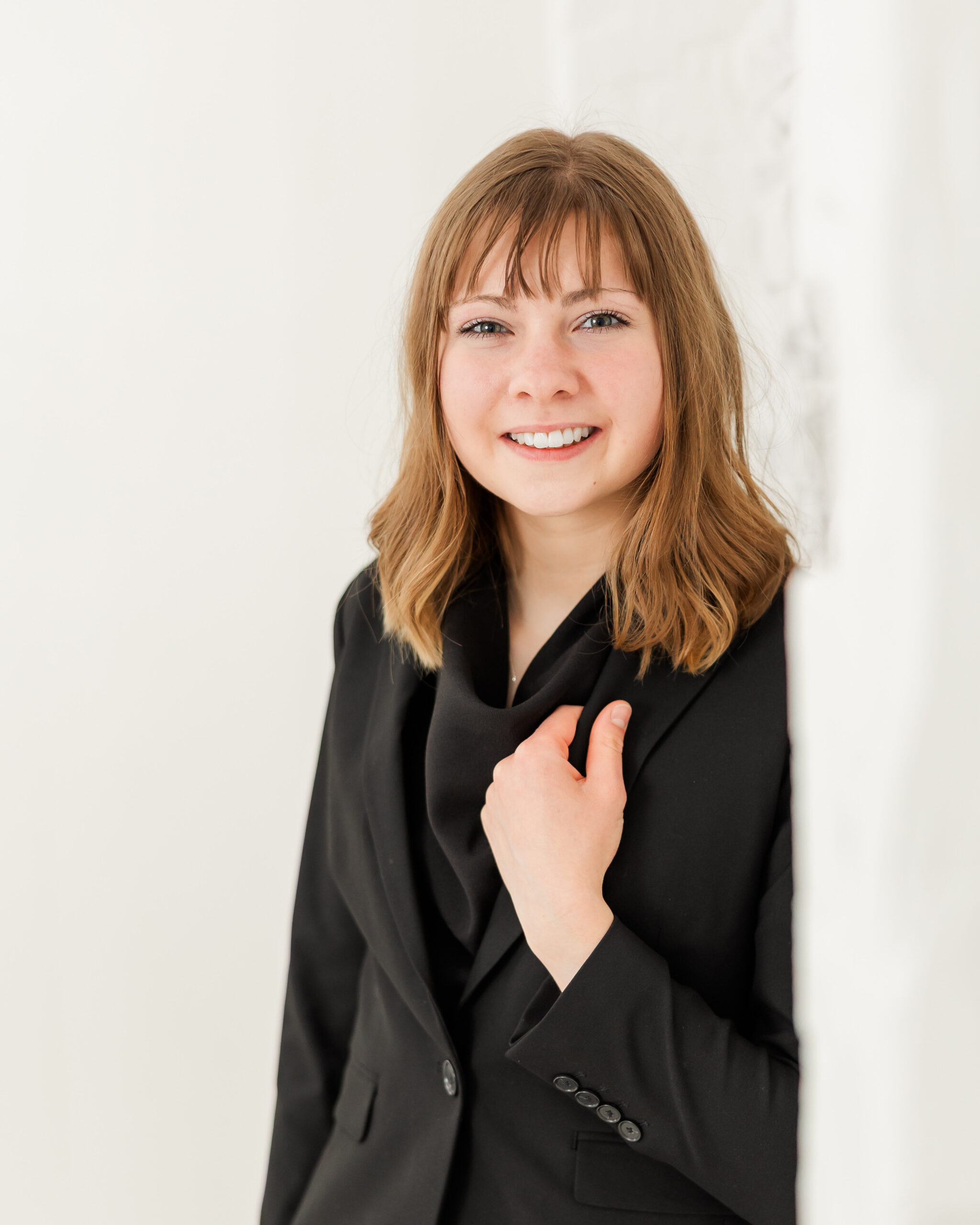 Cadence Ann Hall holding her jacket collar and smiling next to a white wall.